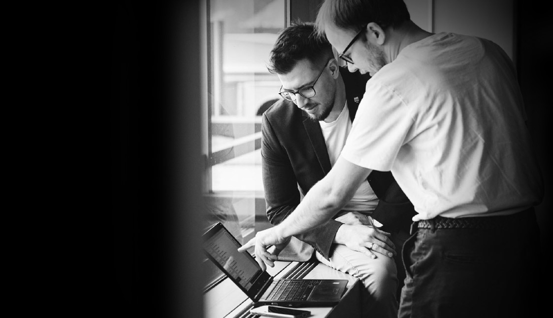 arvenio-b2b-marketing-agentur-rottweil-b2b-marketing-shopdesign-6 Zwei Mitarbeiter sitzend und stehend mit BLick auf einen Laptop