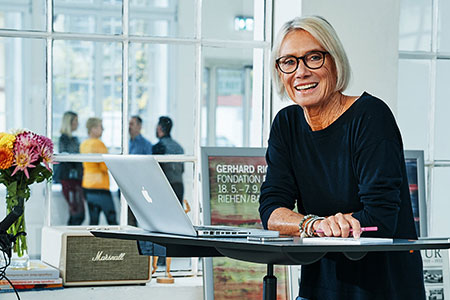 Eine lächelnde Frau an einem Stehpult mit einem Laptop und Kollegen im Hintergrund